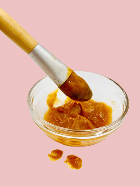 A small clear glass bowl filled with Char's Effective's Pumpkin + Cranberry Enzyme Face Mask which is a thick dark orange creamy mask with a cosmetic brush dipped in the mask against a solid pink background
